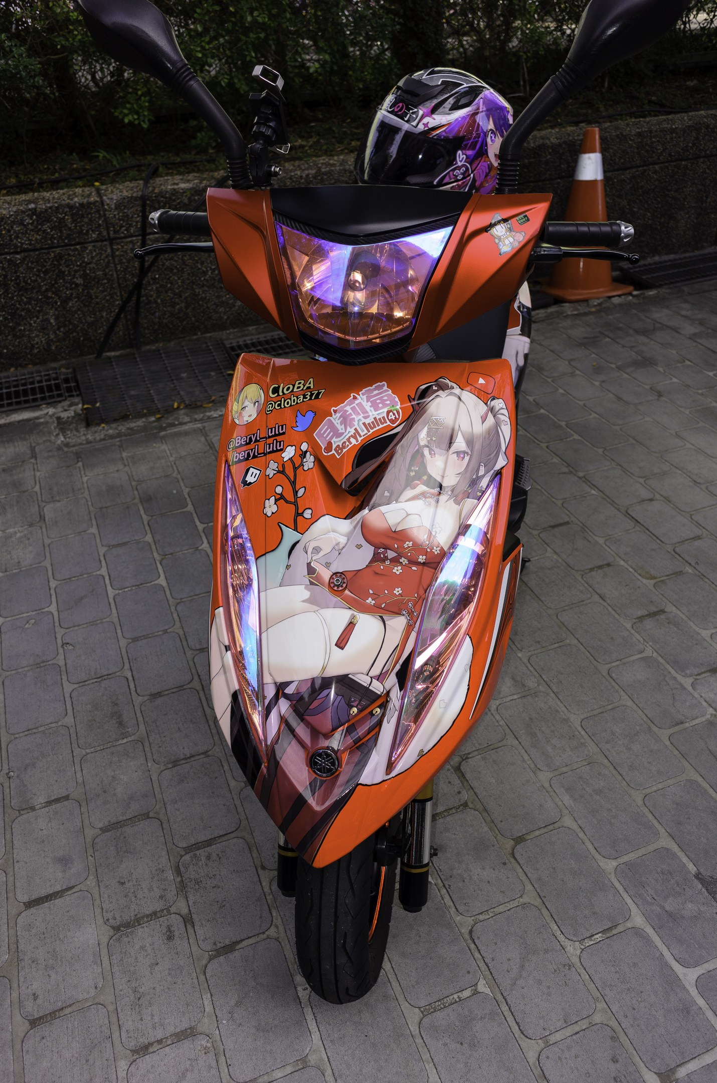 Oranger Roller auf der Straße geparkt mit einem Helm darauf, Verkehrskegel, Wand und Bäume im Hintergrund.