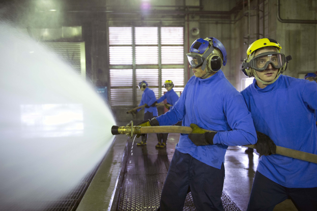 Eine Gruppe von Männern in blauen Hemden und gelben Helmen, die an Maschinen arbeiten, wobei einer Wasser auf den Boden spritzt in einer Fabrik mit sichtbaren Rohren, Fenstern und Lichtern.
