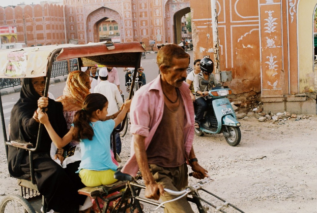 Ein Mann in einem Hemd f├Ąhrt auf einer Rikscha, die zwei Frauen und ein M├Ądchen transportiert, w├Ąhrend ein Mann und eine Frau auf einem Roller nach rechts fahren.