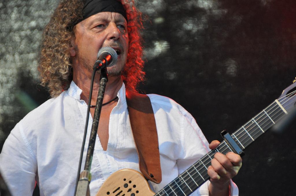 Ein Mann in einem weißen Hemd und einem schwarzen Stirnband singt in ein Mikrofon und hält eine Gitarre.