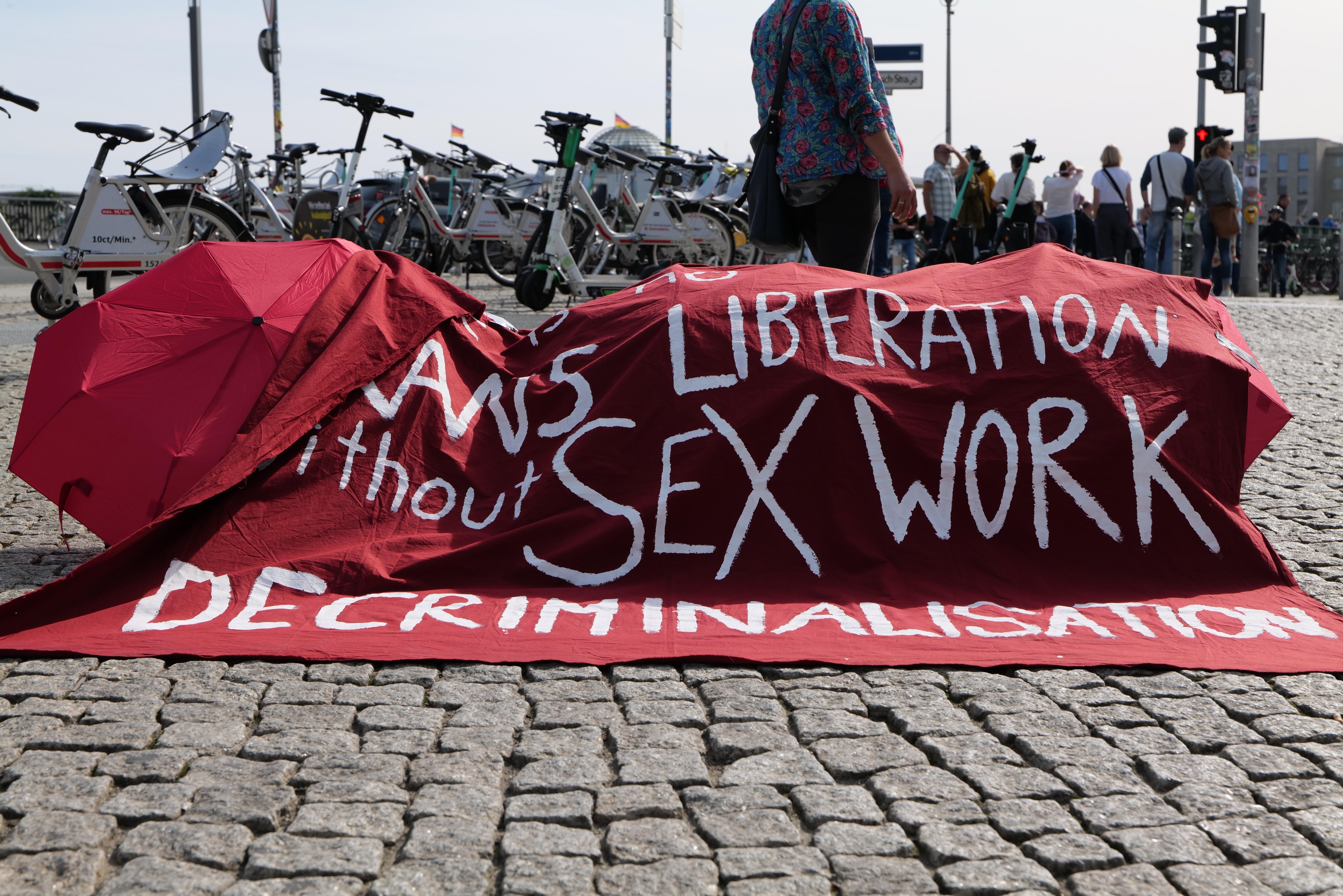 Eine Gruppe von Menschen hält Schirme in der Nähe eines roten Tuchs mit der Aufschrift "Befreiung ohne Entkriminalisierung der Sexarbeit" auf dem Boden, mit Fahrrädern, Pfählen, Verkehrsampeln, Schildern, Gebäuden, Bäumen und dem Himmel im Hintergrund.