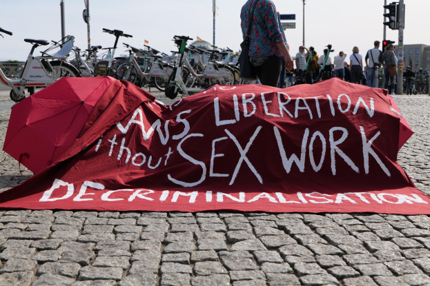 Eine Gruppe von Menschen hält Schirme in der Nähe eines roten Tuchs mit der Aufschrift "Befreiung ohne Entkriminalisierung der Sexarbeit" auf dem Boden, mit Fahrrädern, Pfählen, Verkehrsampeln, Schildern, Gebäuden, Bäumen und dem Himmel im Hintergrund.