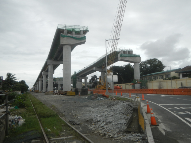 Baustelle mit Brücke im Hintergrund, Straße mit Absperrbaken, verstreute Steine und Gras, Bahnschiene links, Bäume und Gebäude auf beiden Seiten und ein bewölkter Himmel.