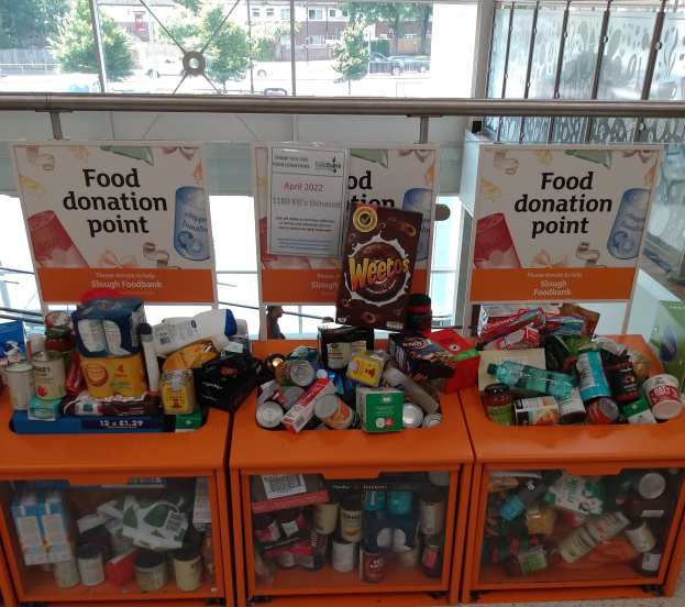 Eine Essensspendestelle mit orangefarbenen Schäubern, die Flaschen, Dosen, Kartons und andere Gegenstände enthalten, begleitet von Informationsschildern, durch Glasfenster betrachtet, die Fahrzeuge, Bäume, Gebäude und Himmel zeigen.