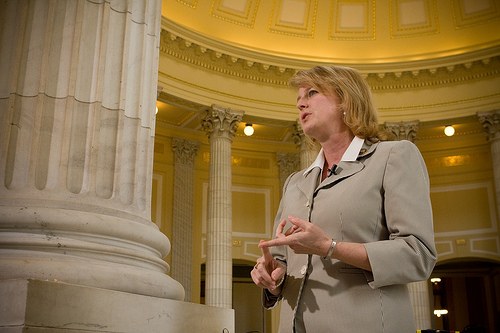 Frau in einem Mantel vor einer beleuchteten Säule in einem Gebäude stehend, die anscheinend eine Politikerin ist.