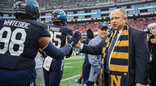 Ein Mann im Anzug und Krawatte gibt einem Fußballspieler die Hand auf einem Feld, umgeben von einer Gruppe von Menschen, mit einer Kamera Person auf der rechten Seite und einem Stadion voller Zuschauer im Hintergrund, das Schilder trägt, die "Notre Dame Football Coach Mike McCarthy und die Fighting Irish" anzeigen.