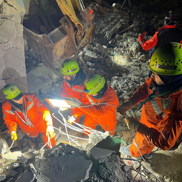 Eine Gruppe von Menschen in orangefarbigen Uniformen und grünen Helmen bei der Arbeit an Schutt von einem eingestürzten Gebäude mit einem Kran im Hintergrund, die Rettungsgeräte halten.