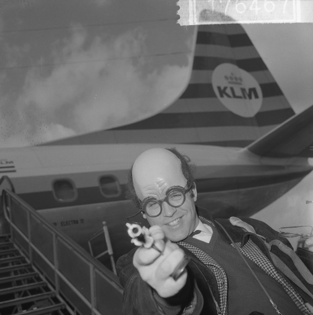 Ein Mann in Anzug und Krawatte hält eine Waffe vor einem großen Flugzeug mit ausgebreiteten Flügeln und brüllenden Motoren, alles in Schwarz-Weiß dargestellt.