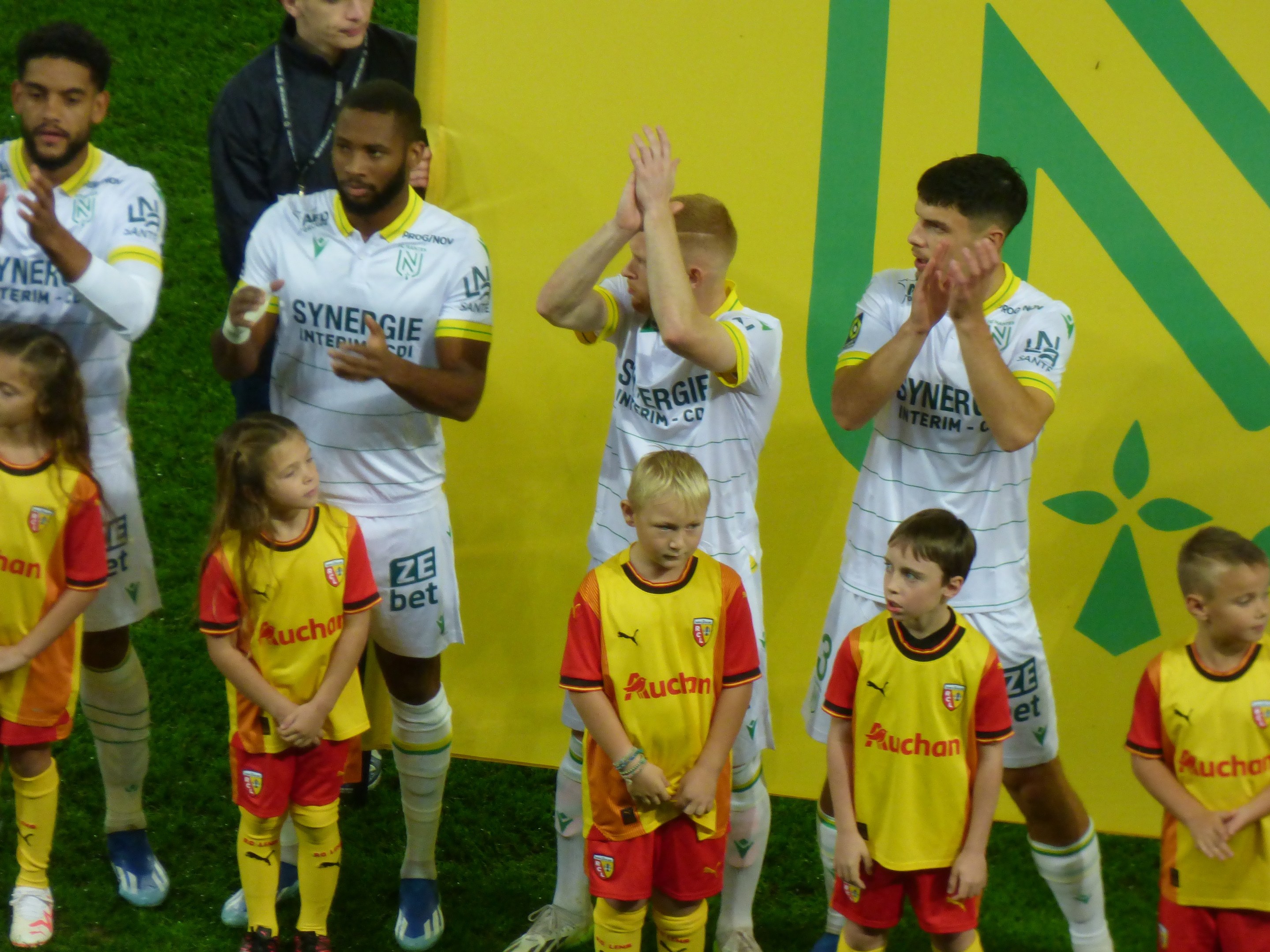 Eine Gruppe von Fußballspielern in Uniformen klatscht auf einem Ligue 1-Feld in Feierlaune, mit einem "Ligue 1" Schild im Hintergrund.