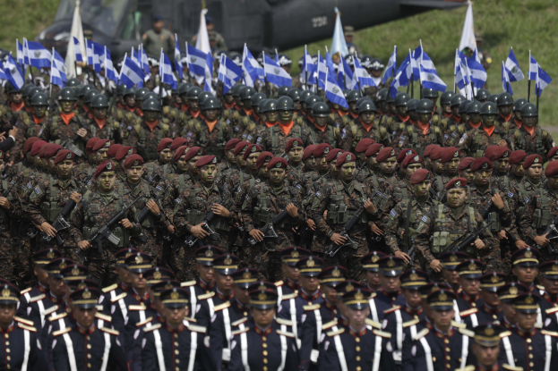 Eine große Gruppe von Soldaten in Mützen und Uniformen marschiert eine Straße entlang und trägt Gewehre und Flaggen bei einer Militärparade mit einem Fahrzeug und Gras im Hintergrund.