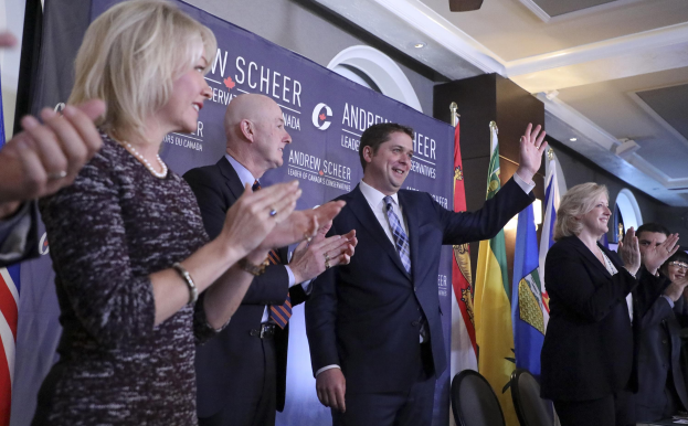 Eine Gruppe von Menschen, die vor Fahnen stehen und klatschen, mit einem Tisch, Stühlen, einer Fahne und einer Wand im Hintergrund.