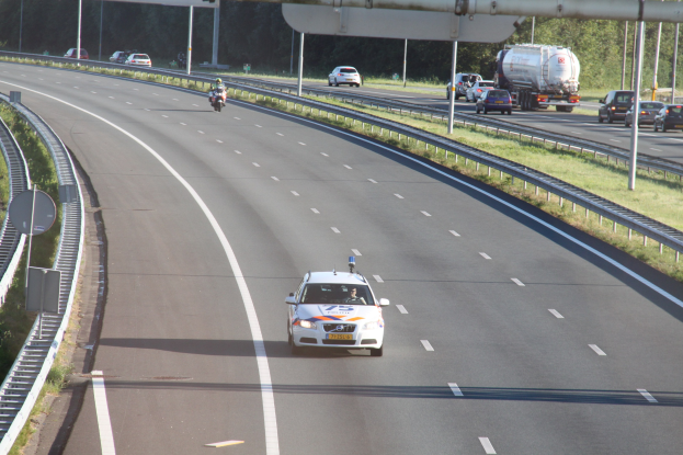 Ein Auto auf einer Autobahn mit einem stationären Polizeiwagen am Rande, andere Fahrzeuge, Gelönder, Pfosten, Schilder und Bäume im Hintergrund.