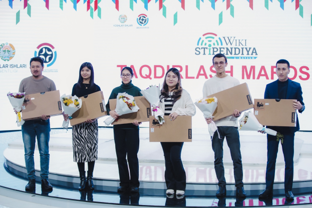 Gruppe von Menschen auf einer Bühne mit Blumensträußen und Pappkartons, im Hintergrund ein Banner mit der Aufschrift "Yoslar Ishlari" und "Wikki Stipendiya"