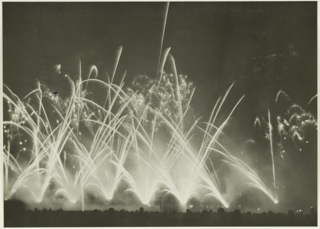 Schwarz-weiß-Foto von Feuerwerk, das in der Nacht explodiert, mit Silhouetten von Menschen am Boden.
