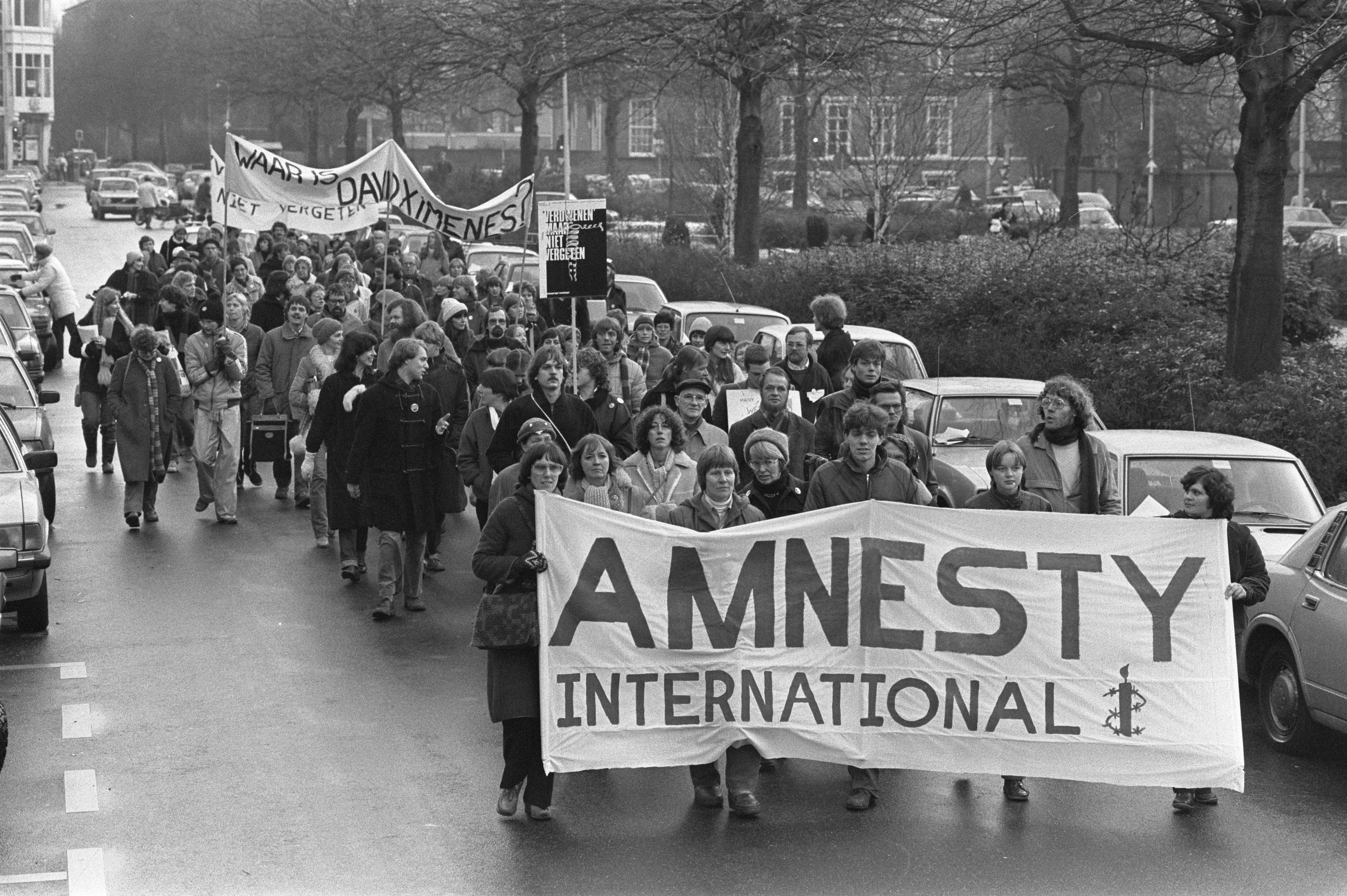 Schwarß-weißes Foto einer Gruppe von Menschen, die eine Straße entlanggehen, mit einer "Amnesty International"-Fahne und parkenden Autos auf beiden Seiten sowie Gebäuden und Bäumen im Hintergrund.