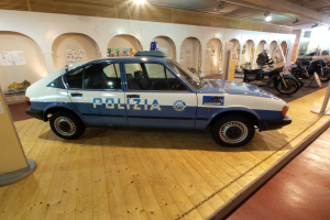 Ein Polizeiwagen in einem Museum ausgestellt, umgeben von Motorrädern und anderen Fahrzeugen, mit einer Säule und einer Informationswand links und Plakaten an den Wänden im Hintergrund.