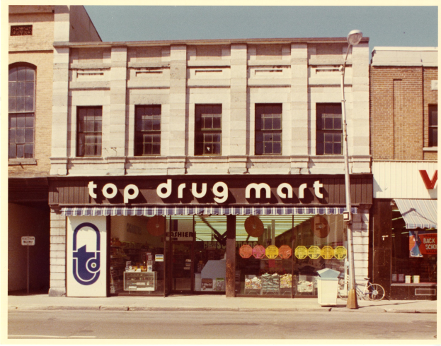 Außenansicht eines Drogstores an einer Straßenecke, mit einem Gebäude mit Fenstern, einer Schildertafel, einem Straßenschild, einem geparkten Fahrrad, einem Müllcontainer und einem bewölkten Himmel.