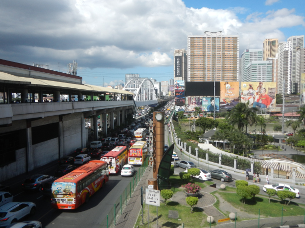 Eine belebte Stra├če mit hohen Geb├Ąuden, Fahrzeugen, einer Brücke mit Passanten, B├Ąumen, Gras, Beschilderung und einem bew├Âlktem Himmel.
