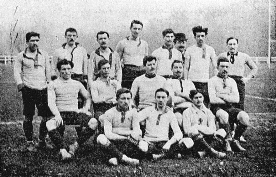 Schwarzes und weißes Foto einer Fußballmannschaft aus den 1920er Jahren mit einigen stehenden und anderen sitzenden Mitgliedern vor Pfählen, Bäumen und einem klaren Himmel.