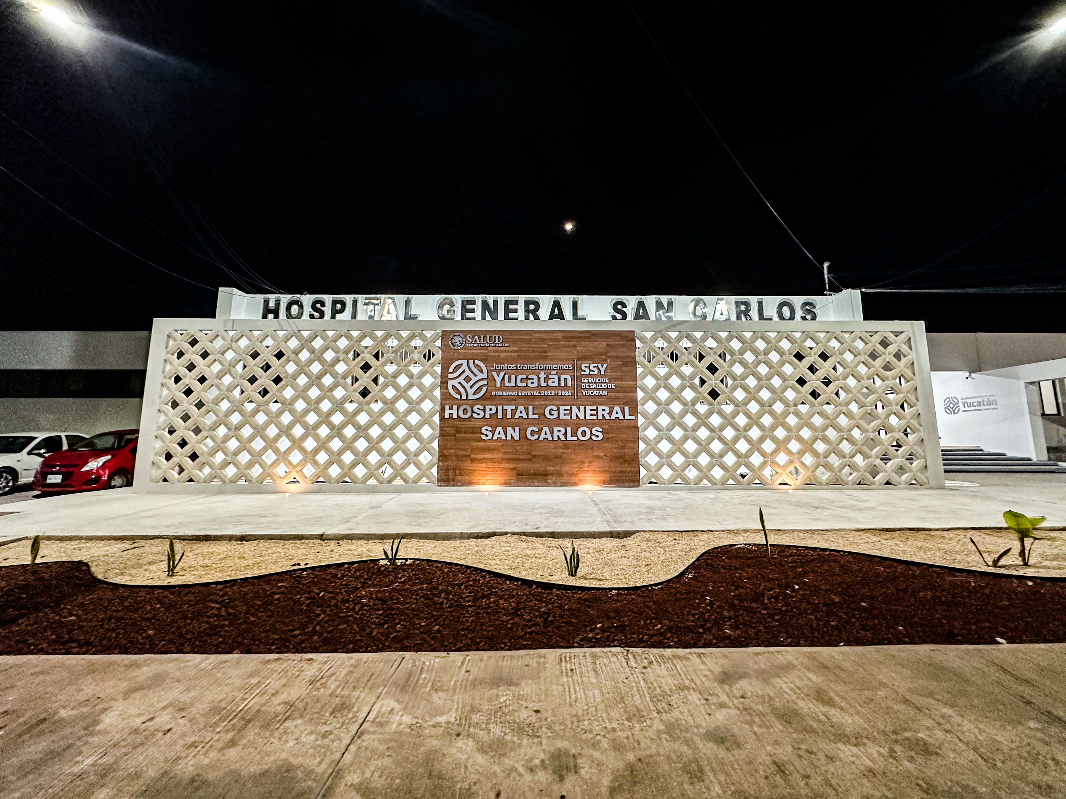 Der Eingang des Hospital General San Carlos hat ein Wandschild, Pflanzen am Boden, geparkte Autos, ein mehrstöckiges Gebäude mit Fenstern, eine Treppe, Lampen, Kabel und einen bewölkten Himmel.