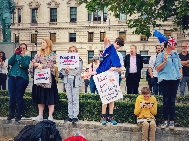 Eine Gruppe von Menschen mit Schildern und Fahnen vor einem Gebäude mit einer Statue auf einem Sockel, umgeben von Pflanzen und Bäumen, während einer Demonstration mit verstreuten Taschen und Gegenständen auf dem Boden.