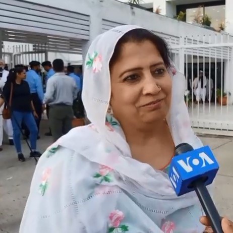 Eine Frau in einem weißen Sari hält ein Mikrofon, während sie zwischen einer Gruppe von Menschen im Freien steht, mit einem Metallzaun, Topfpflanzen, einem Gebäude, einem Baum und dem Himmel im Hintergrund.