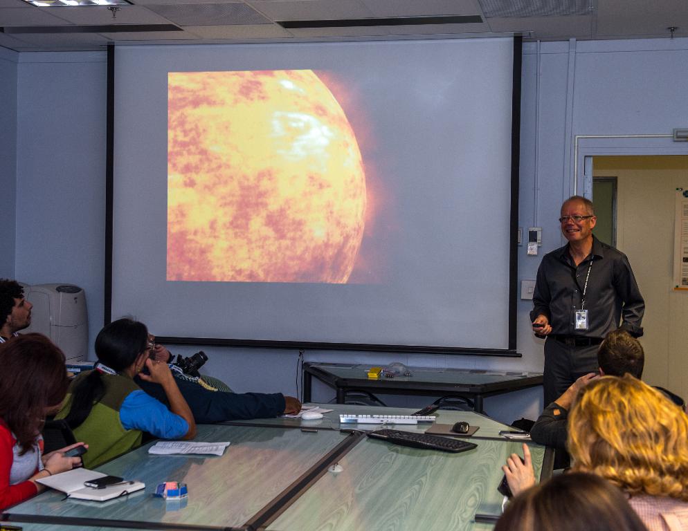 Ein älterer Mann in Hemd und Hose mit einem Ausweis steht rechts und lächelt, während mehrere Personen auf beiden Seiten eines Tisches sitzen und auf einen Projektor in der Mitte schauen.