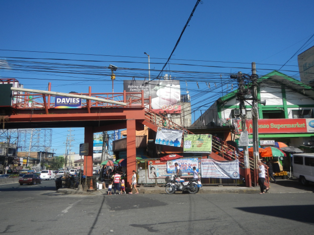 Eine belebte Stadtstraße mit einer Brücke darüber, Fahrzeuge auf der Straße, Fußgänger auf dem Gehweg, Strommasten mit Drähten, Gebäude mit Fenstern, Banner, Straßenlaternen, Bäume und einen klaren blauen Himmel.