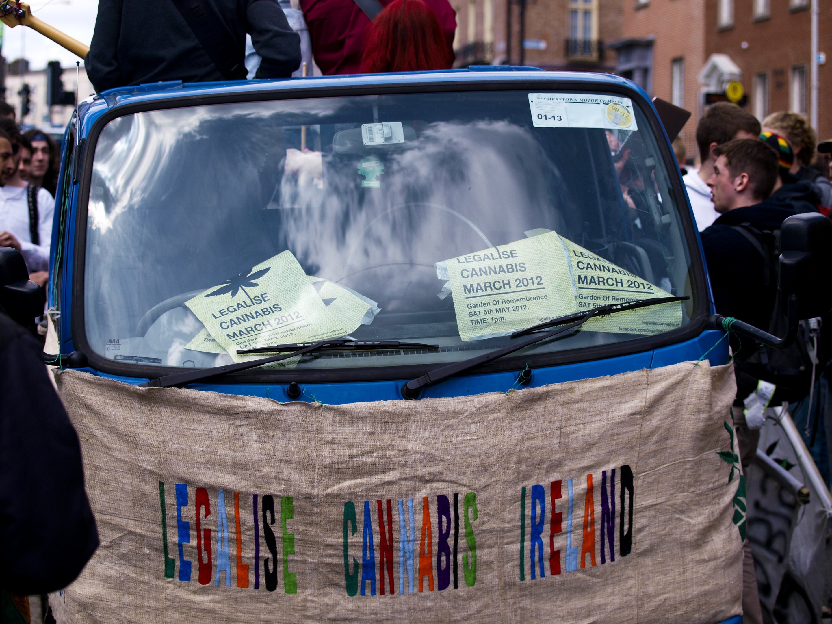 Gruppe von Menschen um ein Auto versammelt mit einem "Legalise Cannabis Ireland"-Schild, Papiere sichtbar im Inneren, vor einem klaren blauen Himmel.