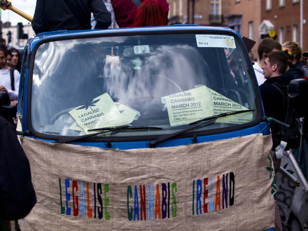 Gruppe von Menschen um ein Auto versammelt mit einem "Legalise Cannabis Ireland"-Schild, Papiere sichtbar im Inneren, vor einem klaren blauen Himmel.
