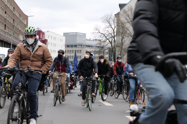 Eine Gruppe von Menschen in Helmen und Handschuhen fährt auf Fahrrädern eine von Bäumen gesäumte Straße in Berlin entlang, mit Gebäuden und einem geparkten Fahrzeug im Hintergrund.
