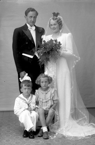 Schwarzes und weißes Foto einer Braut in einem weißen Kleid, die einen Strauß hält, ein strahlender Bräutigam neben ihr und zwei Kinder, die vor ihnen auf dem Boden sitzen.