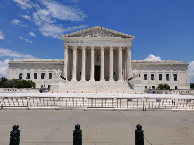 Das Supreme Court-Gebäude in Washington, DC, mit weißen Wänden, Säulen, Treppen und Skulpturen, vor einem Hintergrund aus Bäumen und einem klaren blauen Himmel.