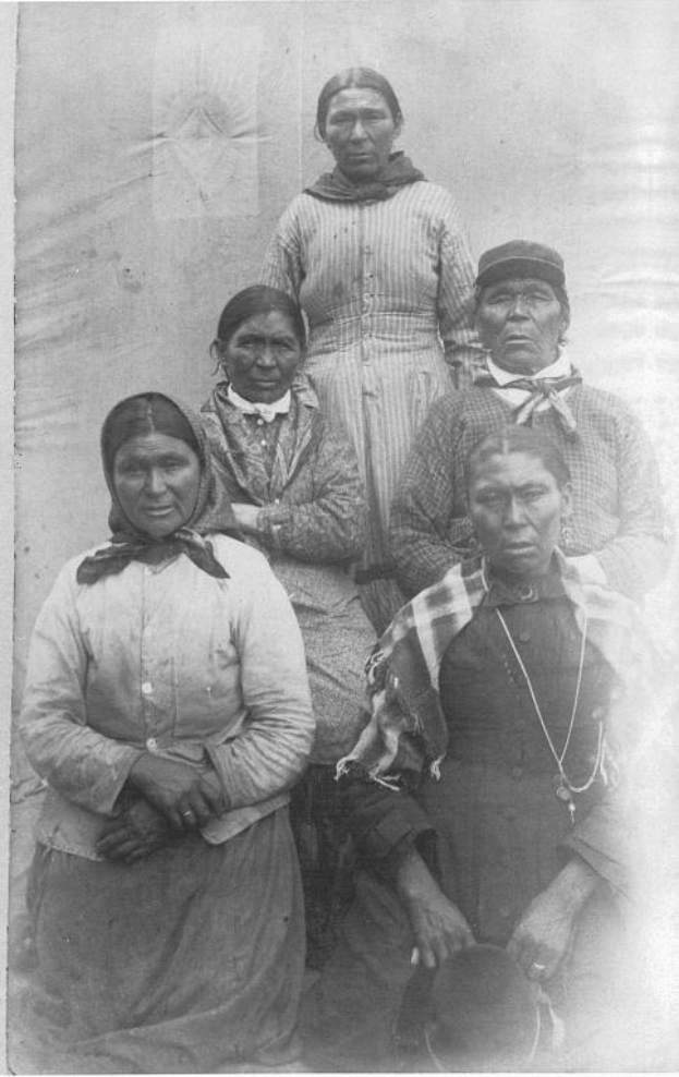 Schwarz-weiß-Fotografie einer Gruppe von indigenen Frauen, die in einer Reihe vor einer schlichten Wand stehen, einige tragen traditionelle Kleidung und Kopfschmuck.