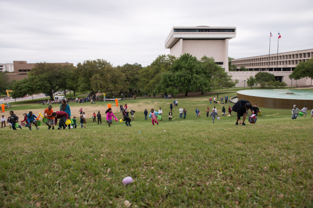 Eine Gruppe von Menschen spielt mit Ostereiern auf einer grünen Wiese mit Bäumen, Gebäuden, Fahnen, einem Brunnen und einem bewölkten Himmel im Hintergrund.