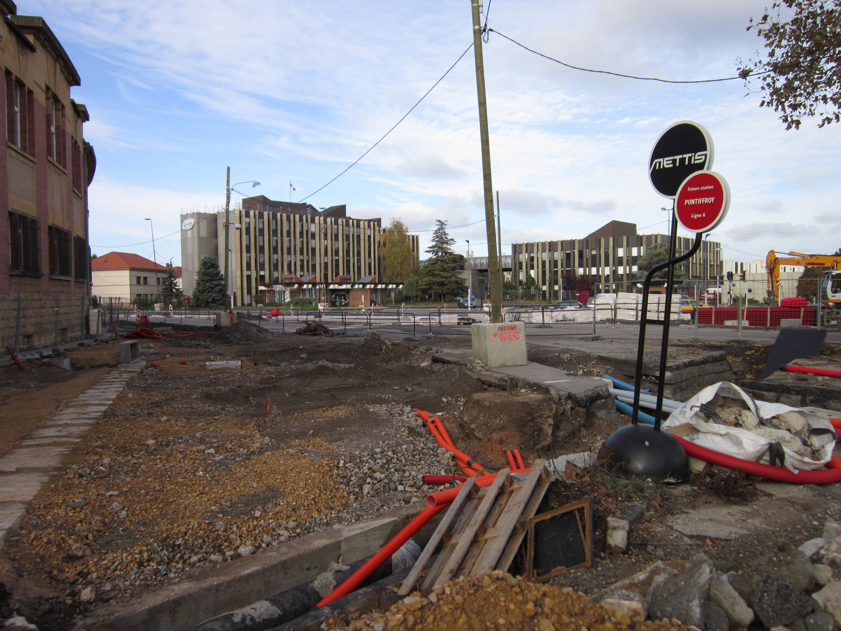 Baustelle mit verstreuten Materialien, Geräten, Hinweistafeln, Absperrungen, Fahrzeugen, Bäumen, Gebäuden, Laternenmasten und einem bewölkten Himmel.