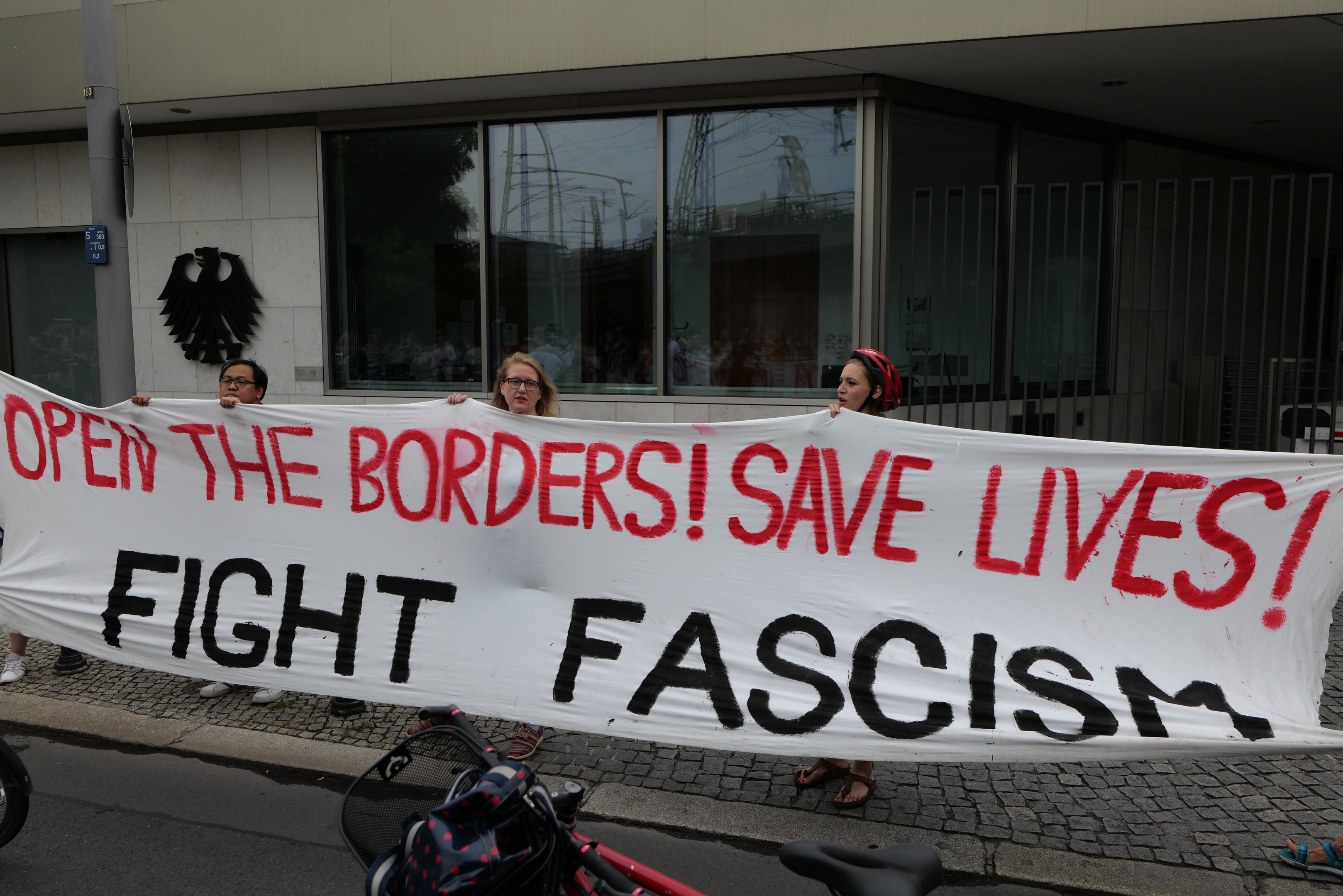 Eine Gruppe von Menschen mit einem Transparent mit der Aufschrift "Öffnet die Grenzen, rettet Leben, kämpft gegen Faschismus" vor einem Gebäude mit Glasfenstern und einem Mast, mit einem Fahrrad und einer Tasche auf der Straße.