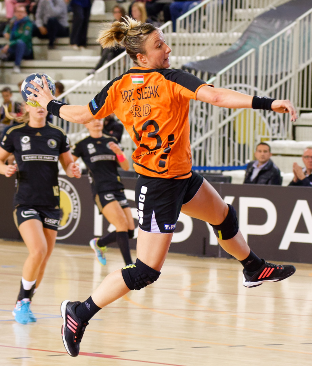 Eine Frau in einem orangen T-Shirt und schwarzen Shorts spielt Handball, hält einen Ball und hat Zuschauer und ein "Krp Slezak - Handball-Weltmeisterschaft 2019"-Schild im Hintergrund.