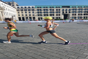 Zwei Frauen in Sportuniformen laufen einen Marathon auf einer Stadtstraße, umgeben von Zuschauern hinter einem Zaun und einer Tafel.