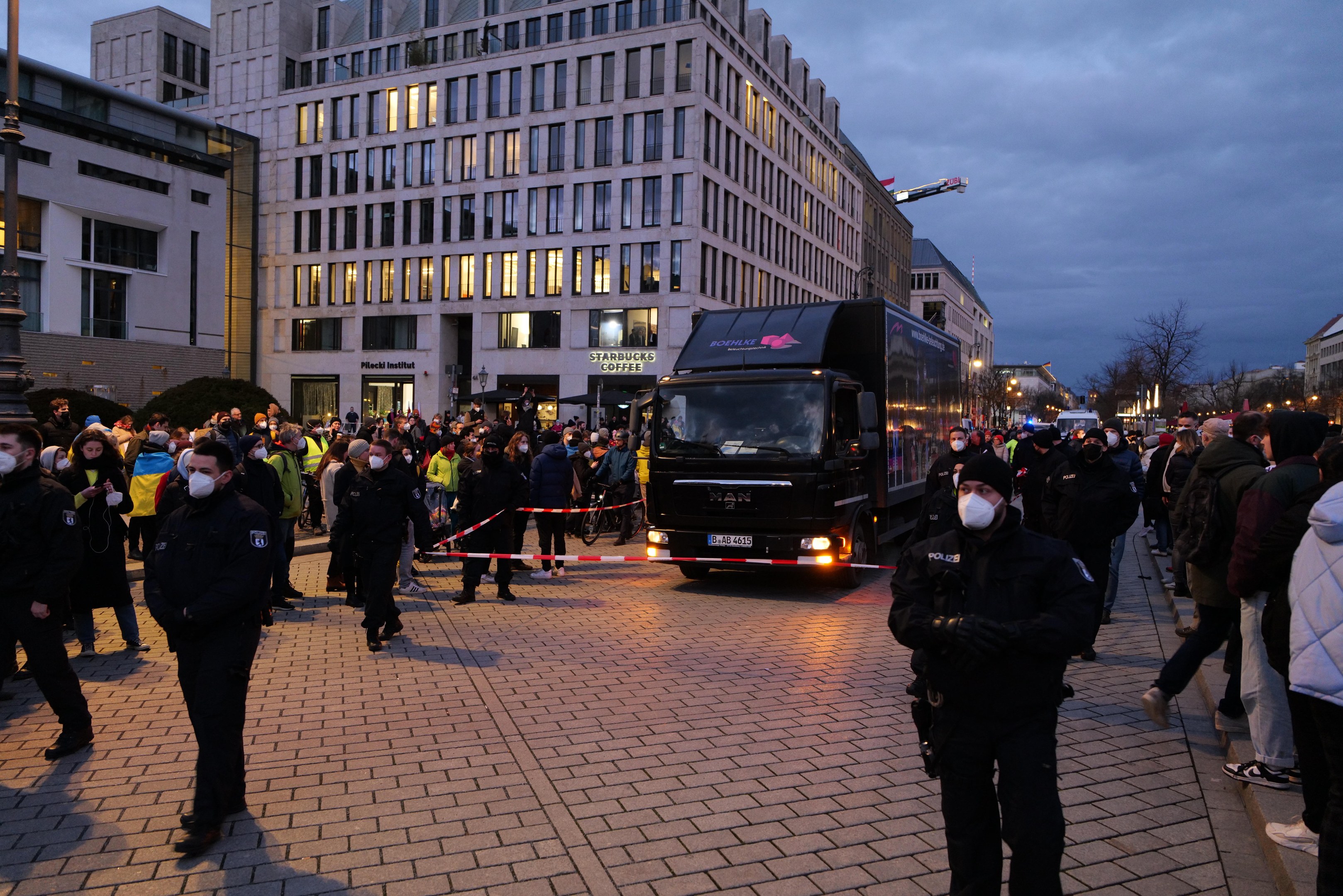 Eine Gruppe von Menschen steht vor einem Lastwagen auf einer Straße, mit Gebäuden, Bäumen und Laternen im Hintergrund, einige tragen Mützen und Masken und ein Band an einem Pfahl im Vordergrund.
