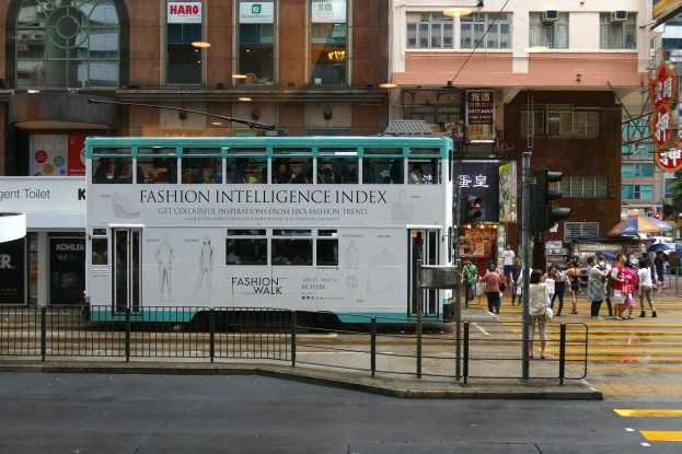 Ein Doppeldeckerbus fährt auf einer Straße mit hohen Gebäuden, Passanten mit Regenschirmen, Verkehrsampeln, einem Geländer, Informationsschildern, Bäumen und einem klaren blauen Himmel.