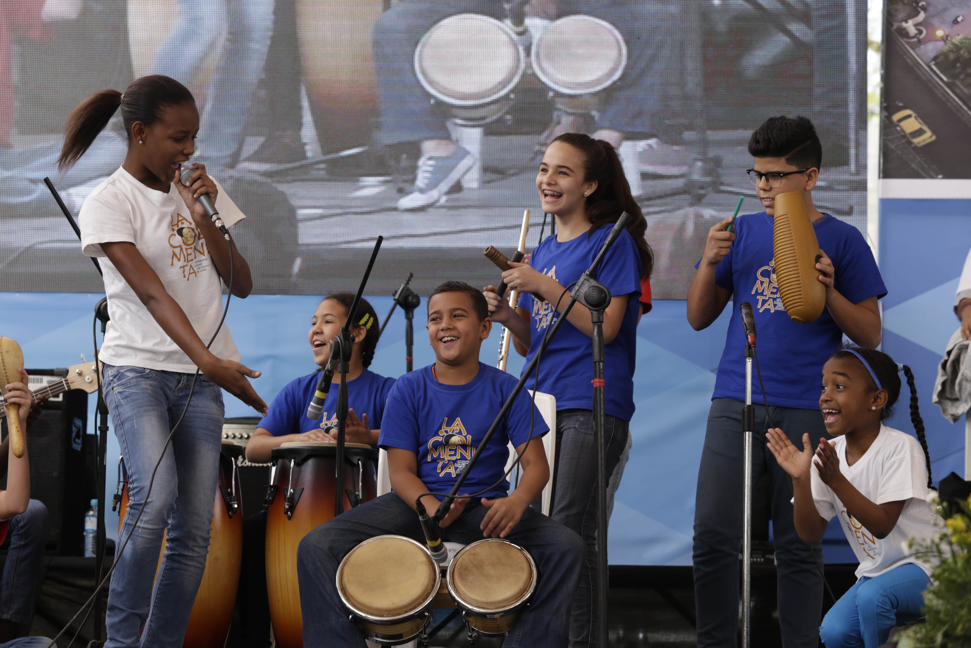 Eine Gruppe von Kindern, die auf einer Bühne Musikinstrumente spielen, mit einer Frau, die ein Mikrofon hält, in der Mitte, umgeben von Mikrofonstativen und einem Banner im Hintergrund, mit Pflanzen auf der rechten Seite.