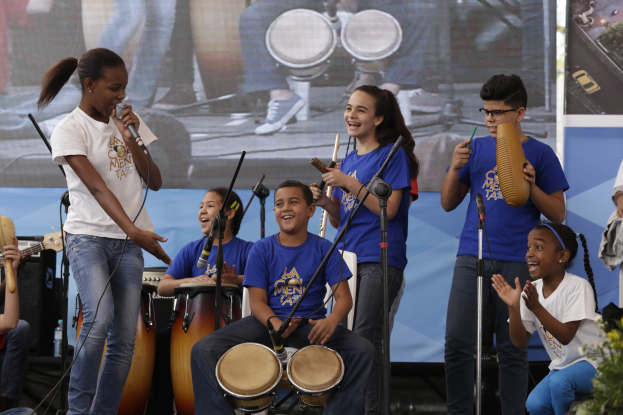 Eine Gruppe von Kindern, die auf einer Bühne Musikinstrumente spielen, mit einer Frau, die ein Mikrofon hält, in der Mitte, umgeben von Mikrofonstativen und einem Banner im Hintergrund, mit Pflanzen auf der rechten Seite.