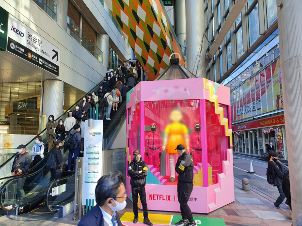 Eine Gruppe von Menschen steht vor einem Gebäude mit Glaswänden und Texttafeln, einige tragen Masken, während eine Rolltreppe mit Menschen daneben fährt; ein pinker Kasten in der Mitte zeigt Schaufensterpuppen und trägt die Aufschrift "Netflix's new Netflix booth in Tokyo."