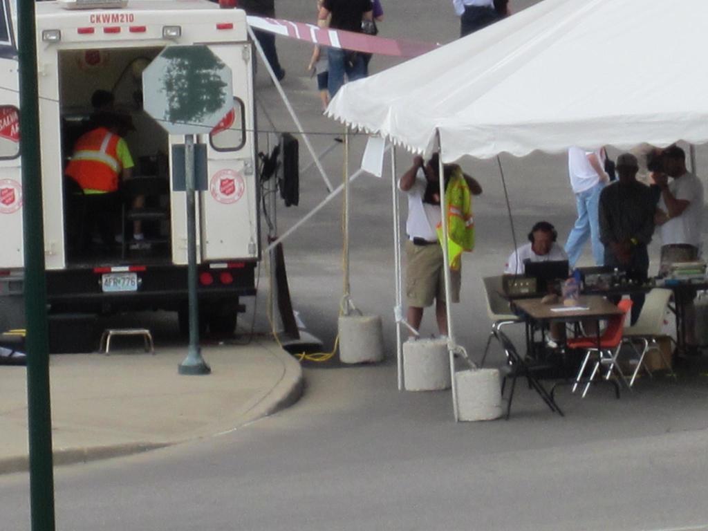 Ein Rettungswagen steht auf der linken Seite einer Stra├če, w├Ąhrend auf der rechten Seite ein Zelt eine Gruppe von Menschen sch├╝tzt, mit einem Tisch und einem Stuhl in der N├Ąhe.