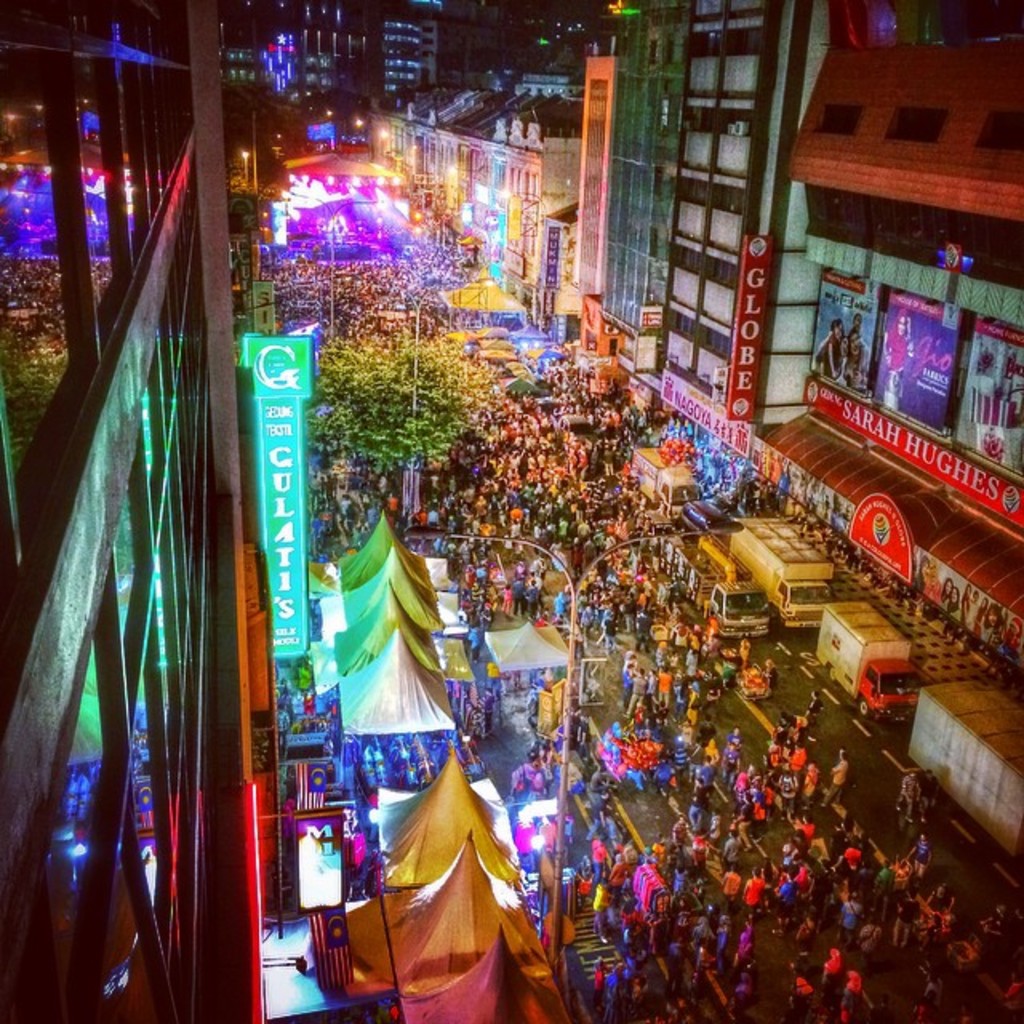 Ein lebhaftes Karnevalsszenario mit einer großen Menschenmenge auf der Straße, umgeben von Zelten, Geschäften, Gebäuden und Bannern, mit zahlreichen Lichtern, die den Hintergrundbereich erhellen.