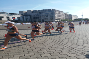Eine Gruppe von Frauen bei einem Marathon auf einer Straße, mit Gebäuden, Bäumen, Polen, Flaggen und einem Turm im Hintergrund unter einem klaren blauen Himmel.
