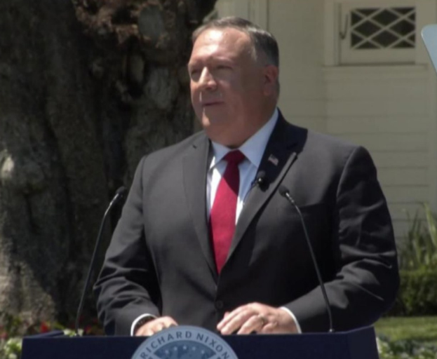 Ein als Staatssekretär Mike Pompeo identifizierter Mann steht an einem Podium mit zwei Mikrofonen und hält eine Rede im Freien in der Nähe eines Baumes und von Pflanzen, mit einem Gebäude im Hintergrund.