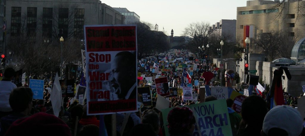 Eine Gruppe von Menschen demonstriert auf einer Straße, hält Plakate hoch, mit Gebäuden, Bäumen und Laternen im Hintergrund unter einem klaren Himmel.