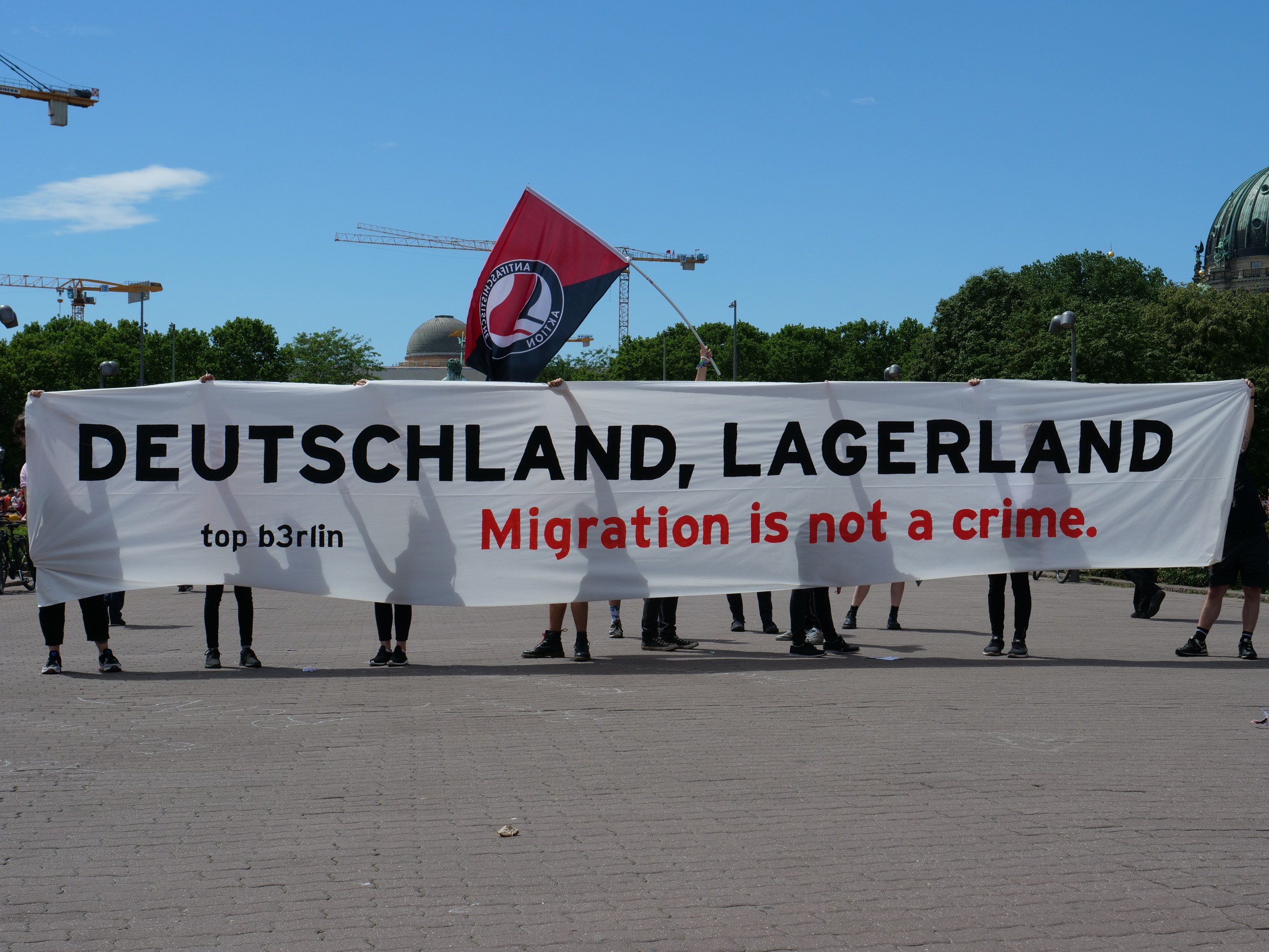 Eine Gruppe von Menschen mit einem Transparent, auf dem "Deutschland, Lagerland Migration ist kein Verbrechen" steht, und einer Flagge, mit Straßenlaternen, Bäumen, Kränen, einem Gebäude und einem bewölkten Himmel im Hintergrund.